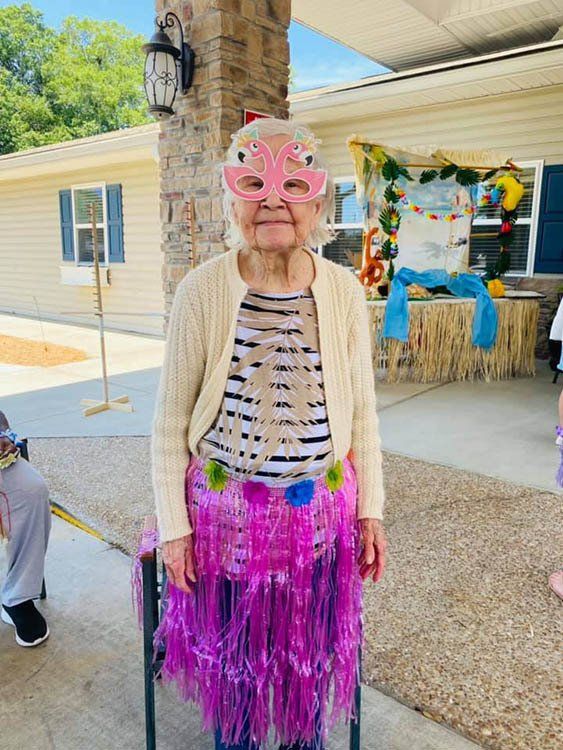 An elderly woman wearing a hula skirt and sunglasses is standing in front of a house.