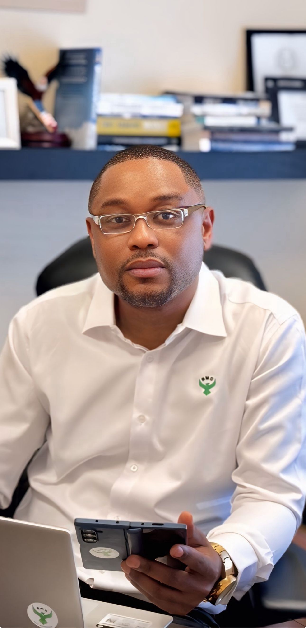 A man in a white shirt is sitting at a desk holding a cell phone.