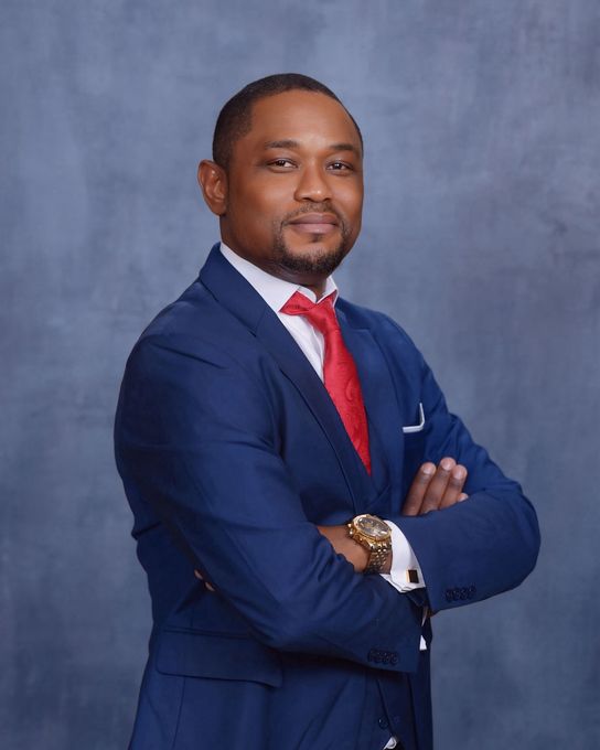 A man in a blue suit and red tie is standing with his arms crossed.