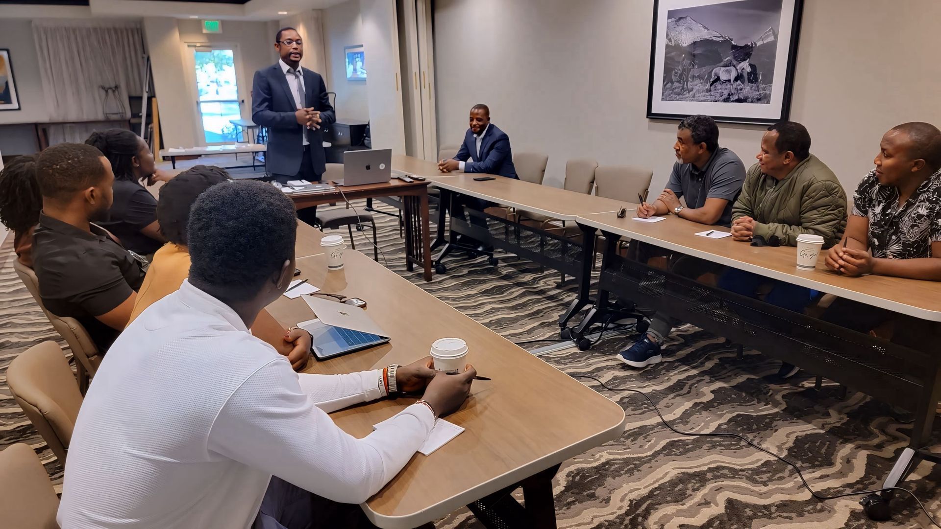 A group of people is sitting at long tables in a conference room.