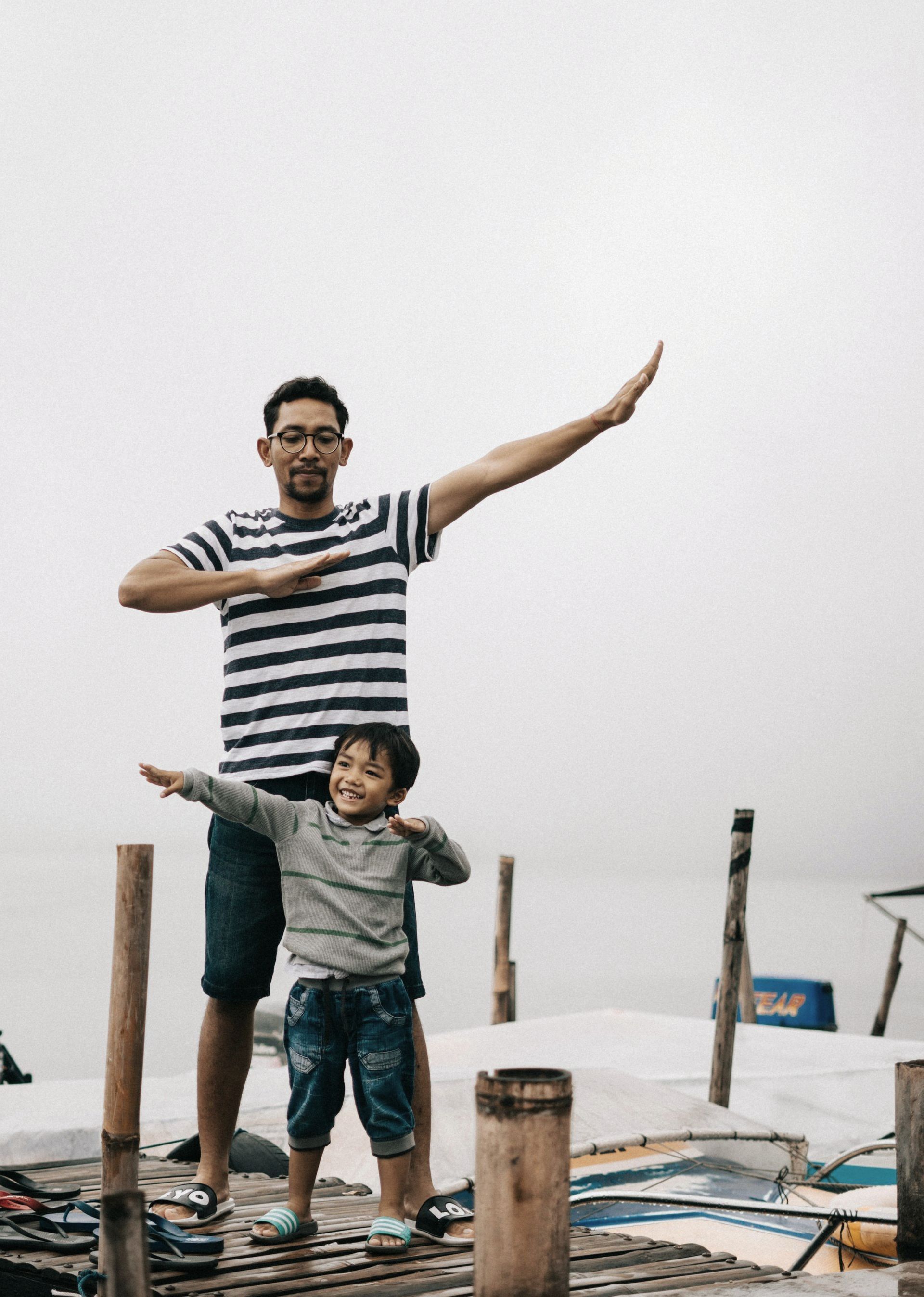A man and a child are standing on a dock with their arms outstretched.