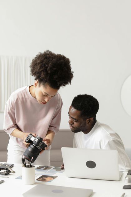 A man and a woman are looking at a camera while sitting at a table with a laptop.