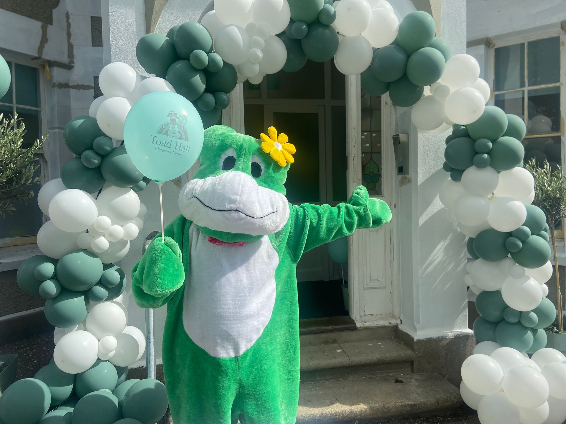 A green frog mascot is holding two balloons in front of a balloon arch.
