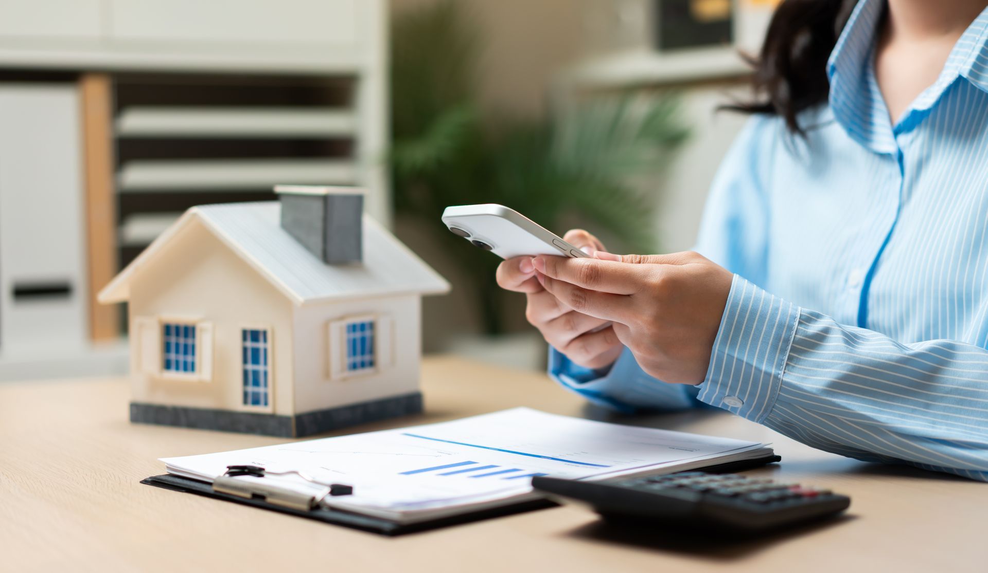 Person holding a smartphone over a desk with a small house model, clipboard with charts, and calculator Person holding a smartphone over a desk with a small house model, clipboard with charts, and calculator