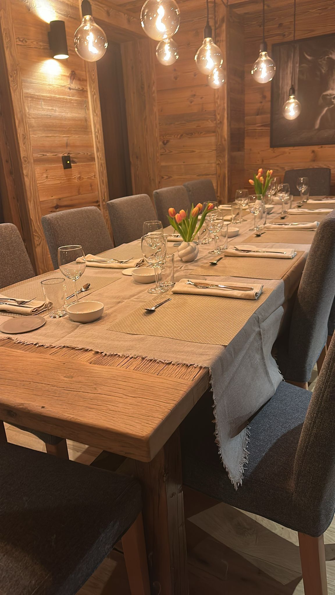 A long wooden dining table set for a formal meal with grey chairs, rustic wood walls, and hanging pendant bulbs.
