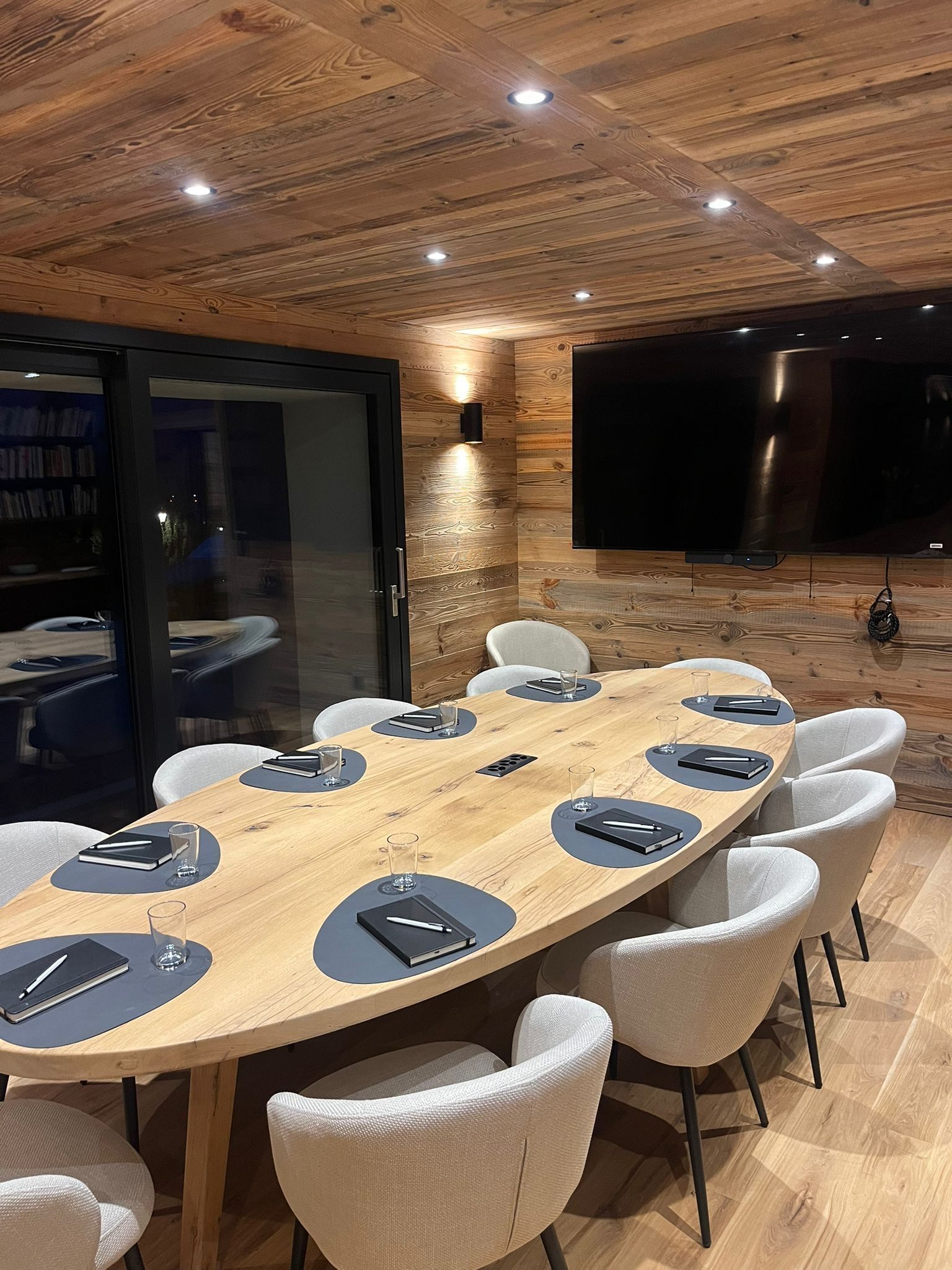 A long oval wooden conference table with grey place mats and white chairs in a rustic room with wood-paneled walls.
