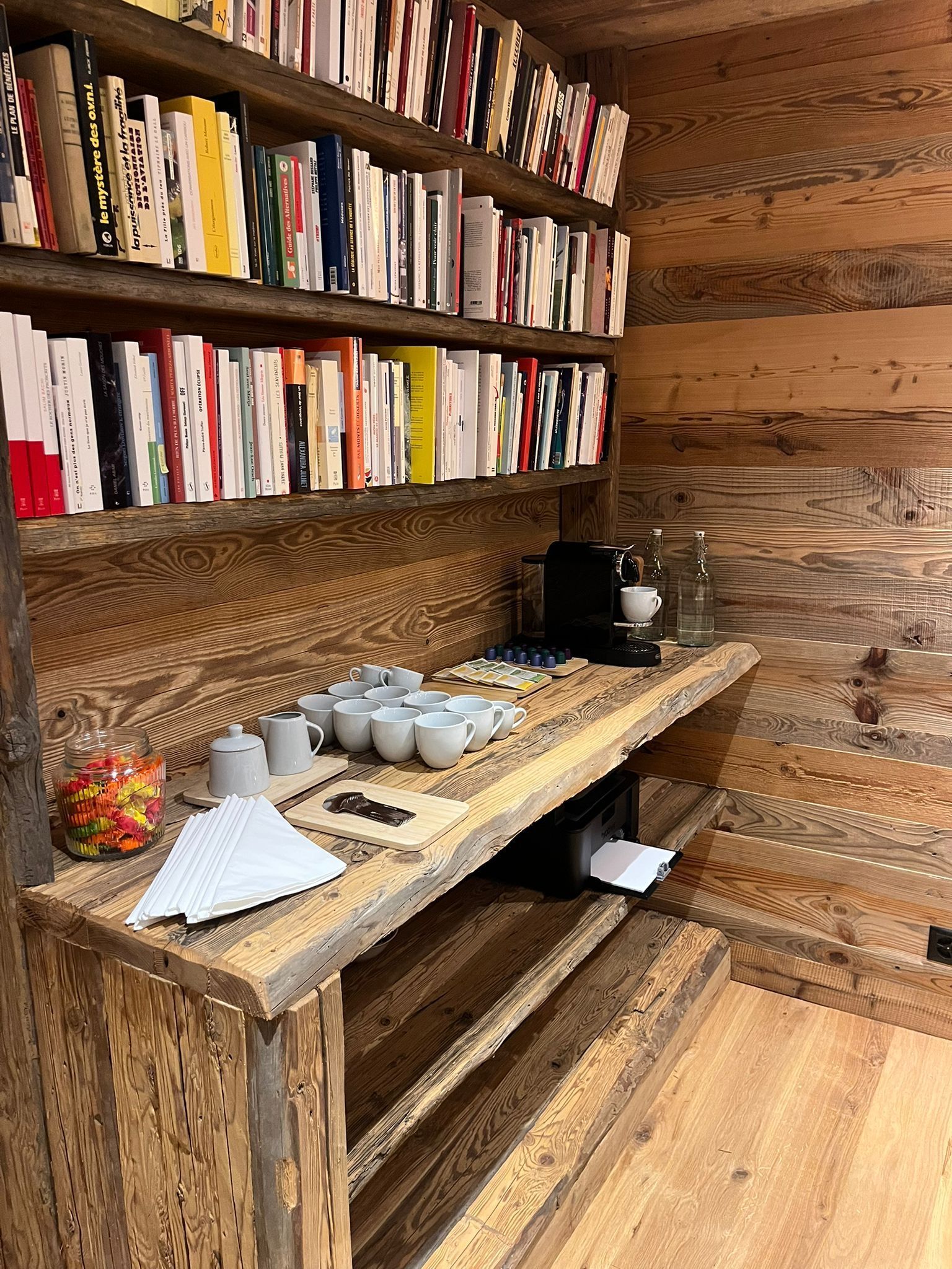 A rustic wooden coffee station with shelves of books, a coffee machine, and small white cups on a matching wood counter.