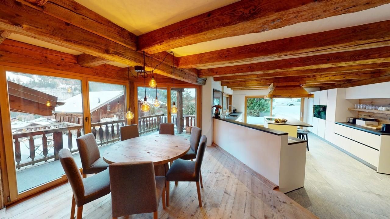 Un espace salle à manger et cuisine au style rustique-moderne, avec un plafond en bois, une table ronde en bois, des chaises grises et une vue sur un paysage enneigé.