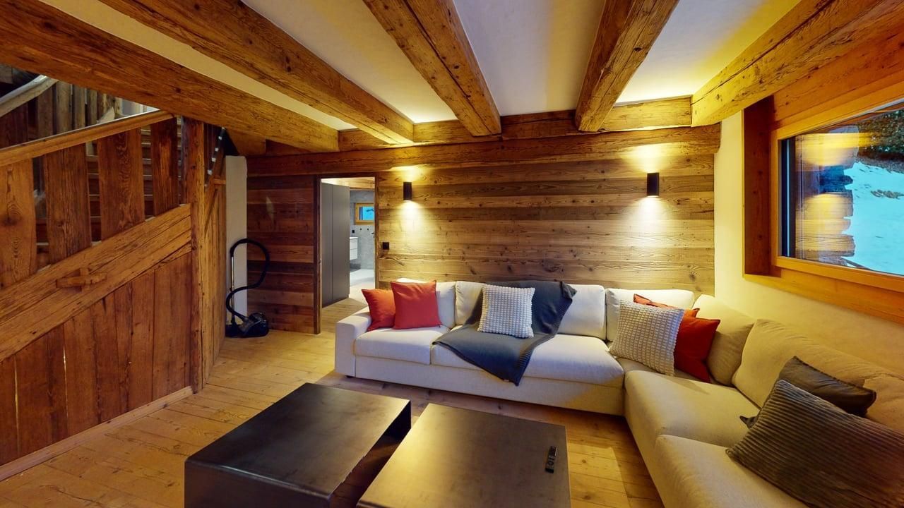 Un salon chaleureux avec des murs en bois, des poutres apparentes au plafond, un canapé d'angle blanc et deux tables basses foncées.