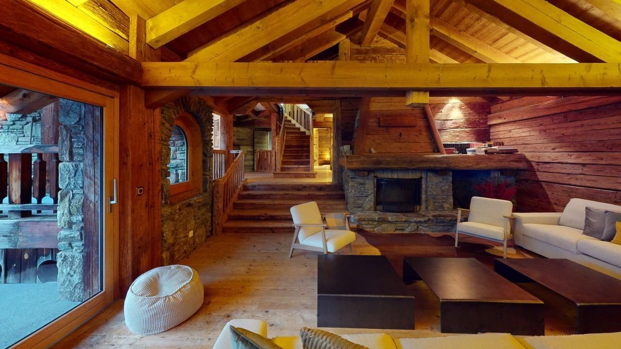 Un salon rustique avec des poutres apparentes, une cheminée en pierre, des fauteuils beiges et un escalier menant à une mezzanine.