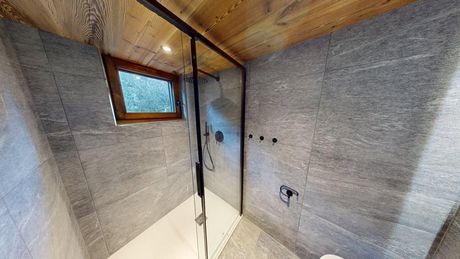 Une douche moderne avec des carreaux de pierre grise, une porte en verre et un plafond en bois, agrémentée d'une petite fenêtre et d'accessoires noirs.