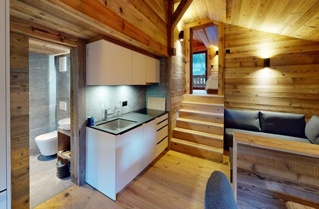 Petite kitchenette avec placards blancs, lambris, canapé gris et escalier menant à une salle de bains et à l'étage.