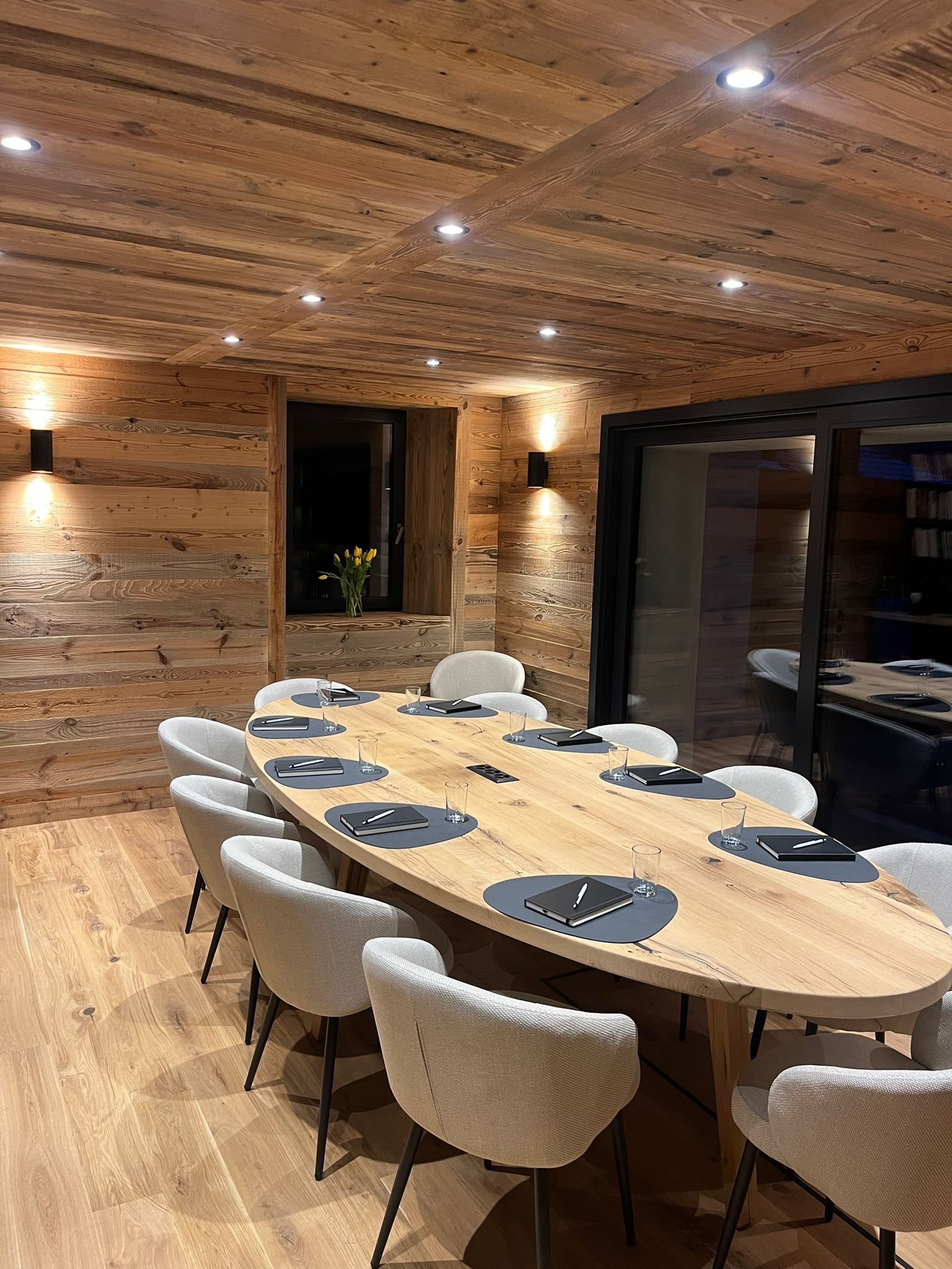 Une salle de conférence moderne, lambrissée, avec une table ovale en bois et des chaises blanches sous un éclairage encastré.