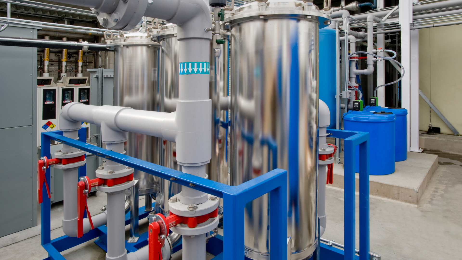 A large stainless steel tank is surrounded by blue pipes in a factory.