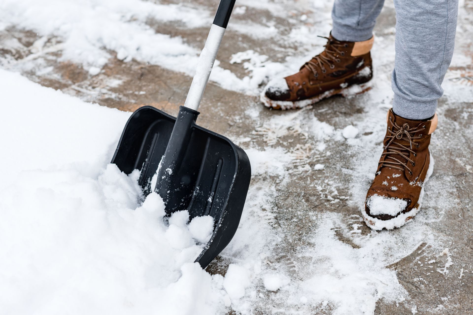 Person shoveling snow off a sidewalk with a black shovel; wearing brown boots and gray pants.