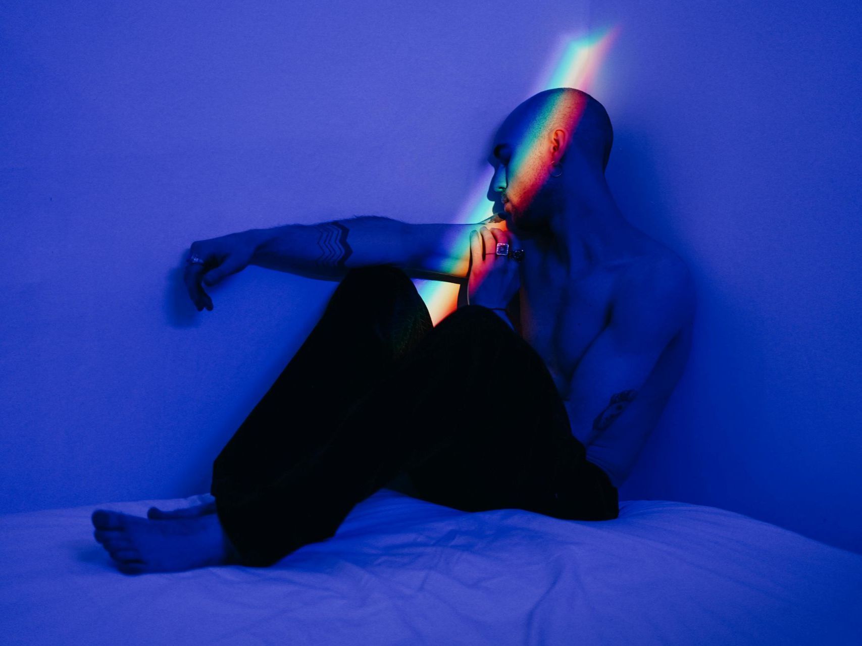 A contemplative gay man sits shirtless in blue lighting, with a rainbow prism across hi