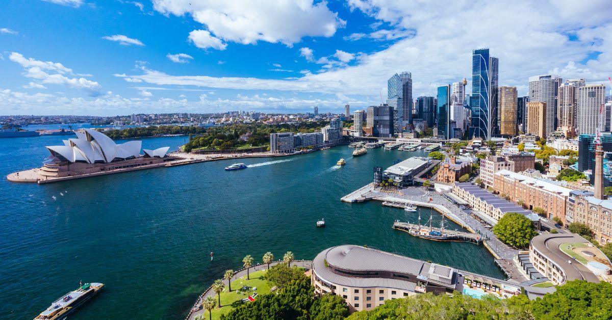 Aerial view of Sydney Harbour featuring the Opera House, ferries, and the city skyline under a bright blue sky