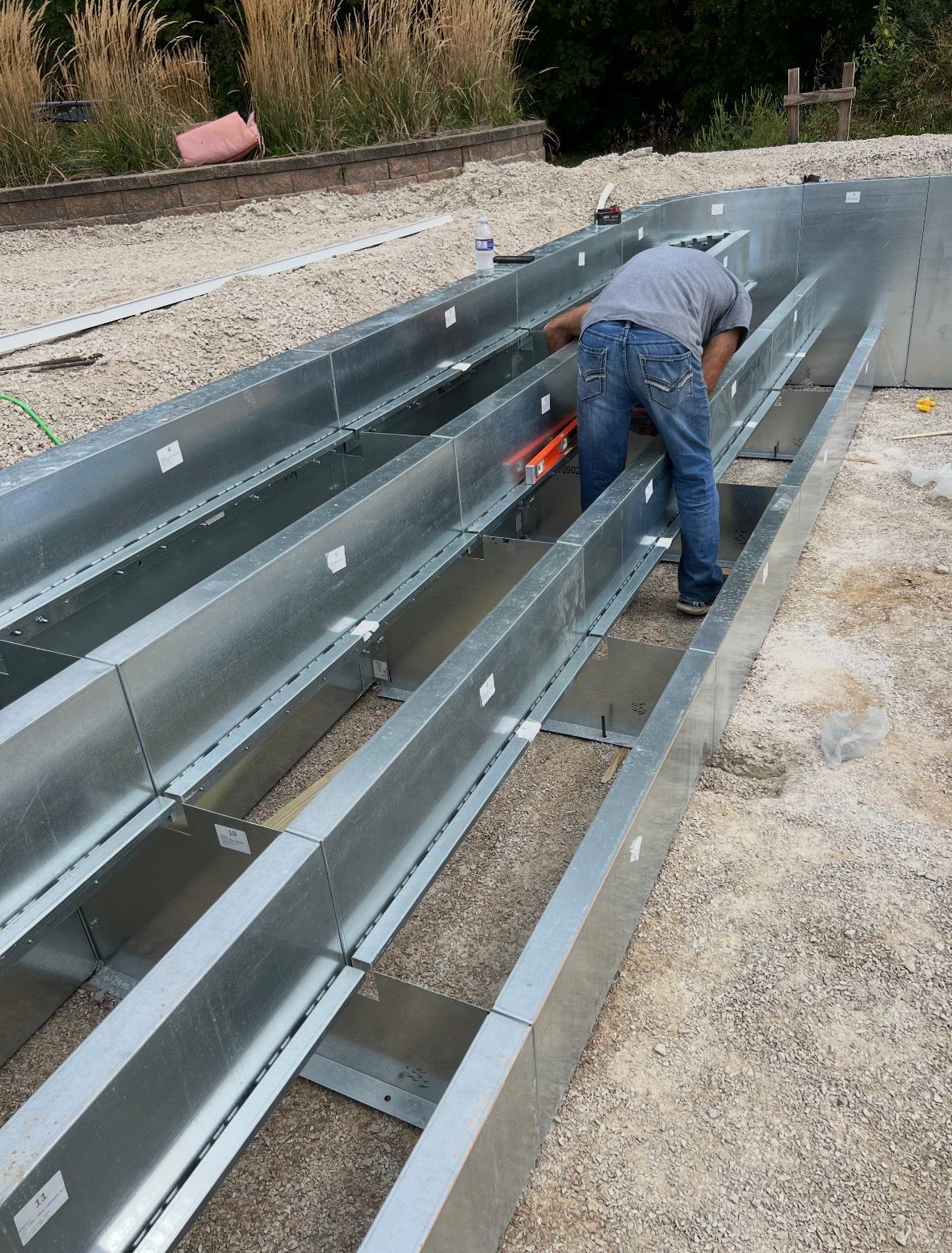 A person in a gray shirt and blue jeans works on a frame made of long, silver metal panels set on a gravel surface.