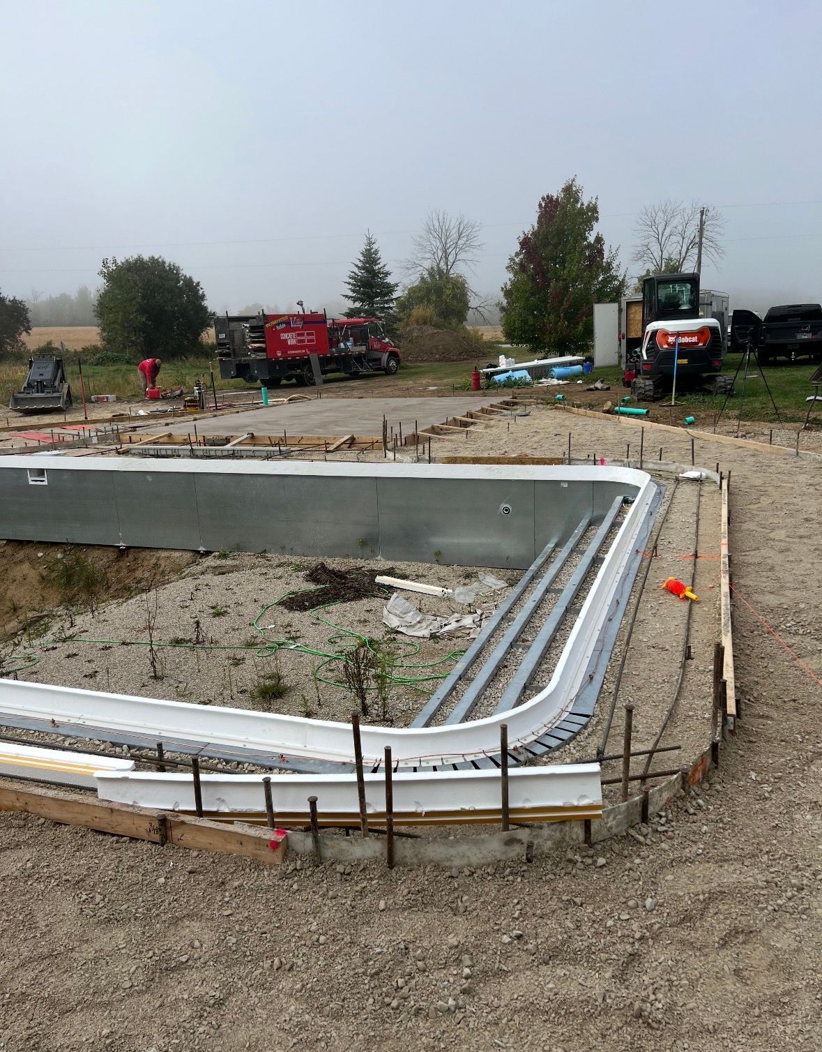 Construction site showing a half-built swimming pool frame with metal walls, white coping, and gravel, with machinery nearby.