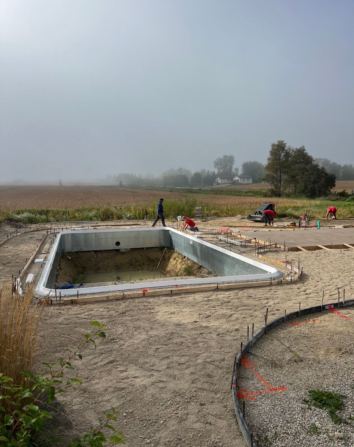 A rectangular, in-ground swimming pool shell under construction in an open, misty field with workers visible nearby.