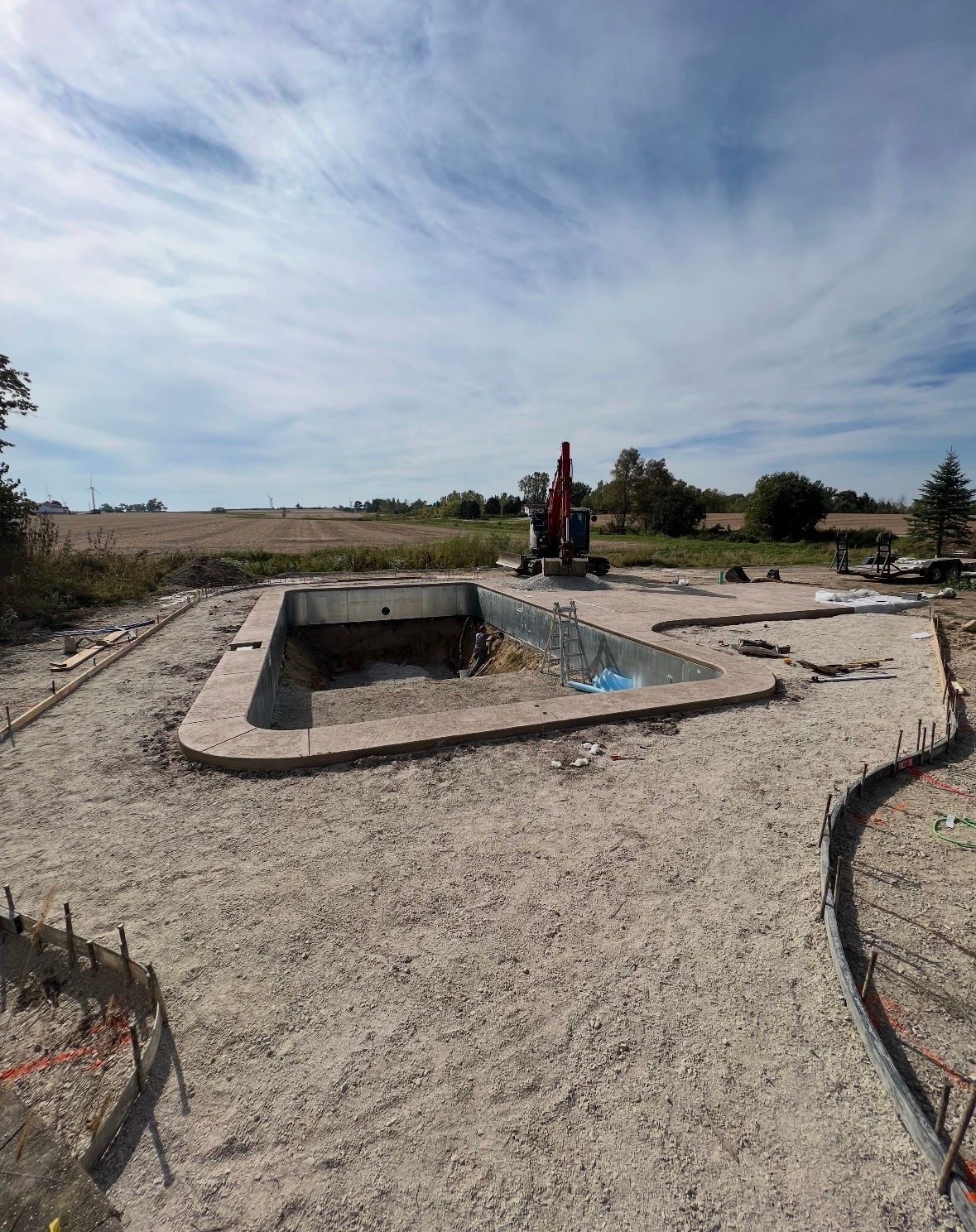 An excavated swimming pool frame sits in a gravel yard with a construction vehicle in the background.
