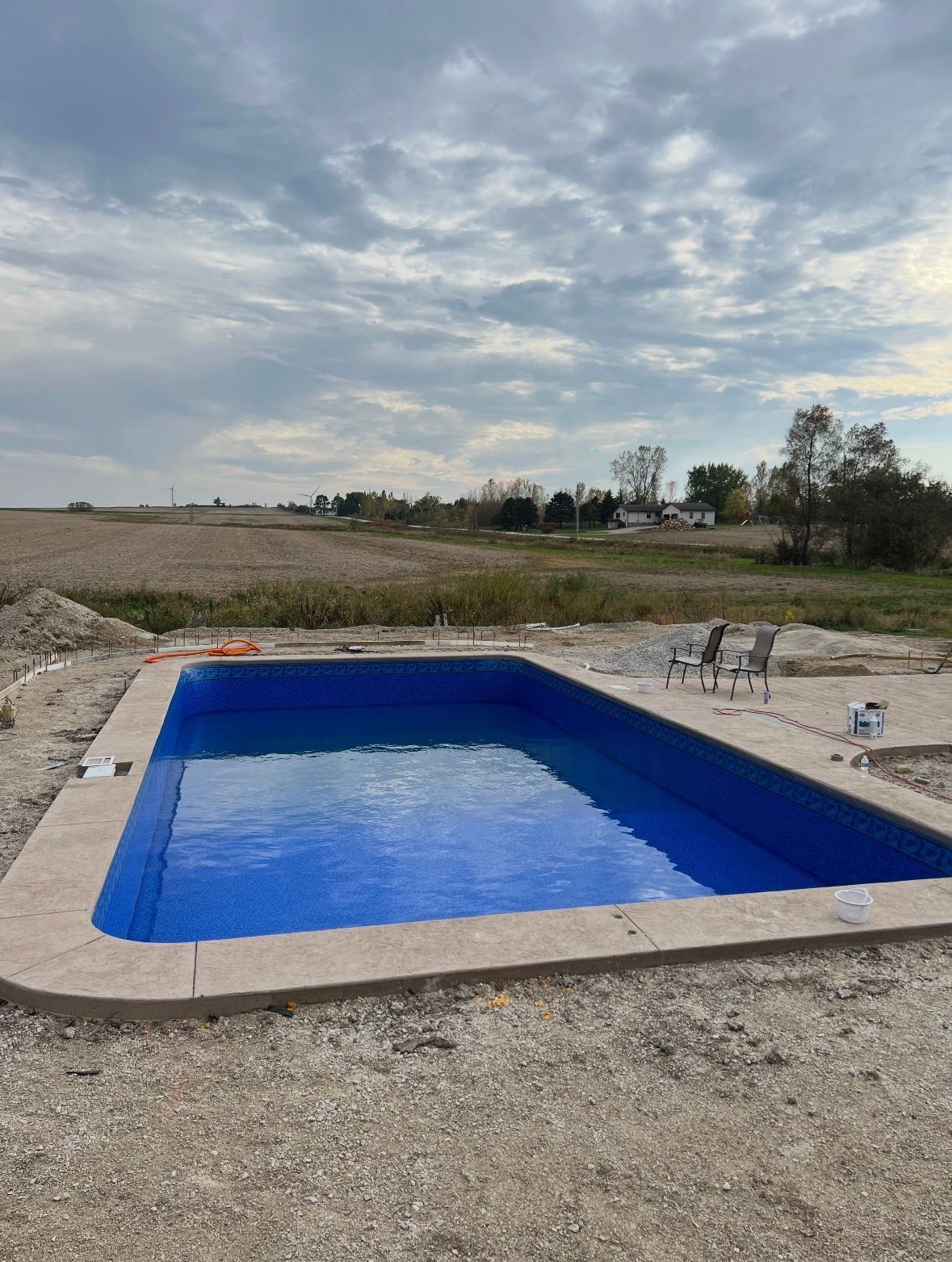 A rectangular, bright blue swimming pool sits in an unfinished gravel yard under a cloudy sky.