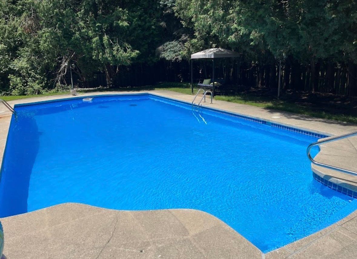 A rectangular blue swimming pool set in a backyard surrounded by lush green trees and a shaded patio area.