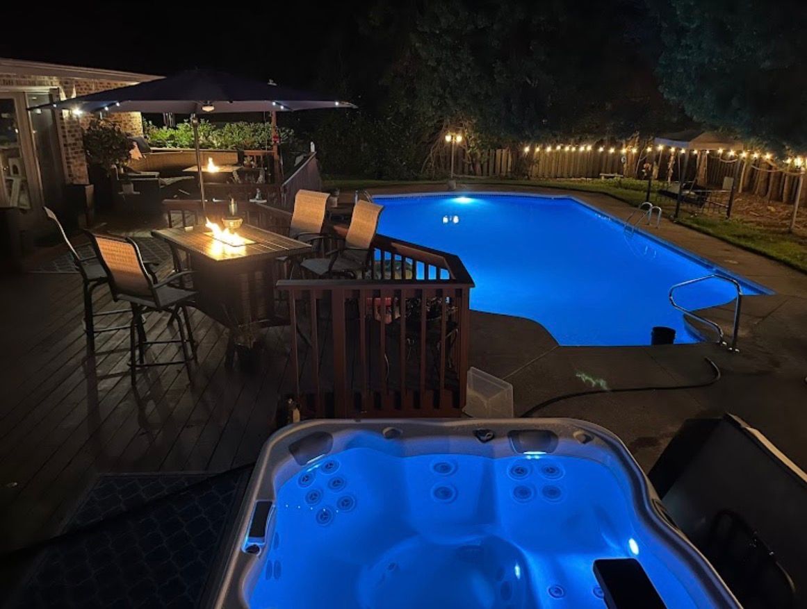 A backyard at night featuring an illuminated hot tub in the foreground, a swimming pool, a fire pit, and string lights.