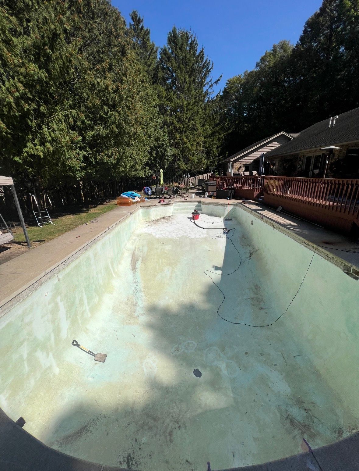 An empty, light-colored rectangular swimming pool surrounded by a patio, wooden deck, trees, and a clear blue sky.