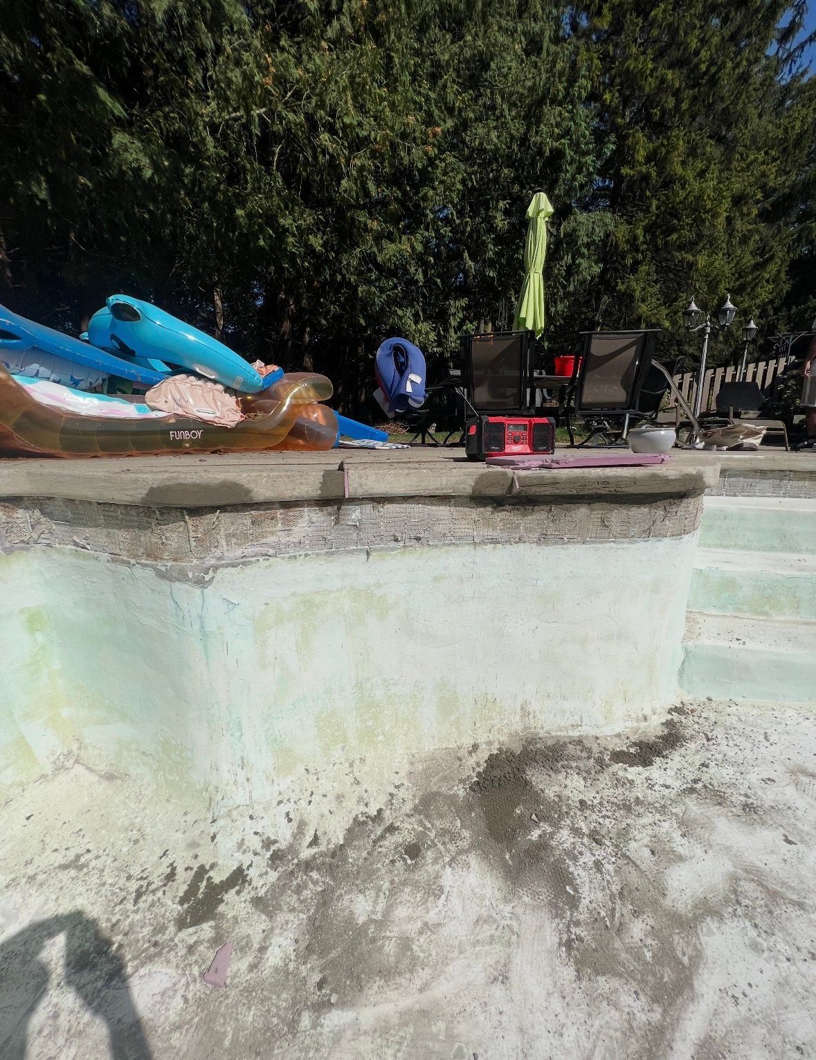 A drained swimming pool with a weathered concrete edge, pool floats on the deck, and dense trees in the background.