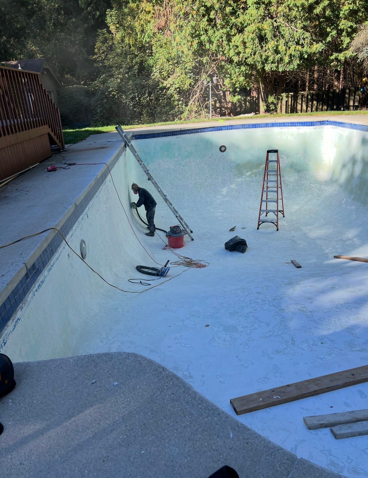 A worker repairs a residential swimming pool, using a ladder and tools while standing inside the empty basin.