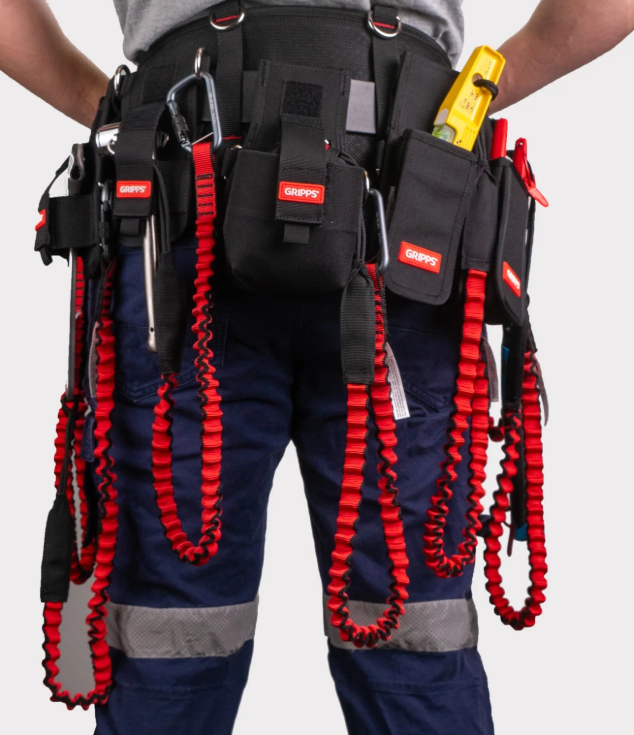 A close-up view of a person wearing a black work belt with several pouches, red and black coiled tool lanyards, and blue work trousers