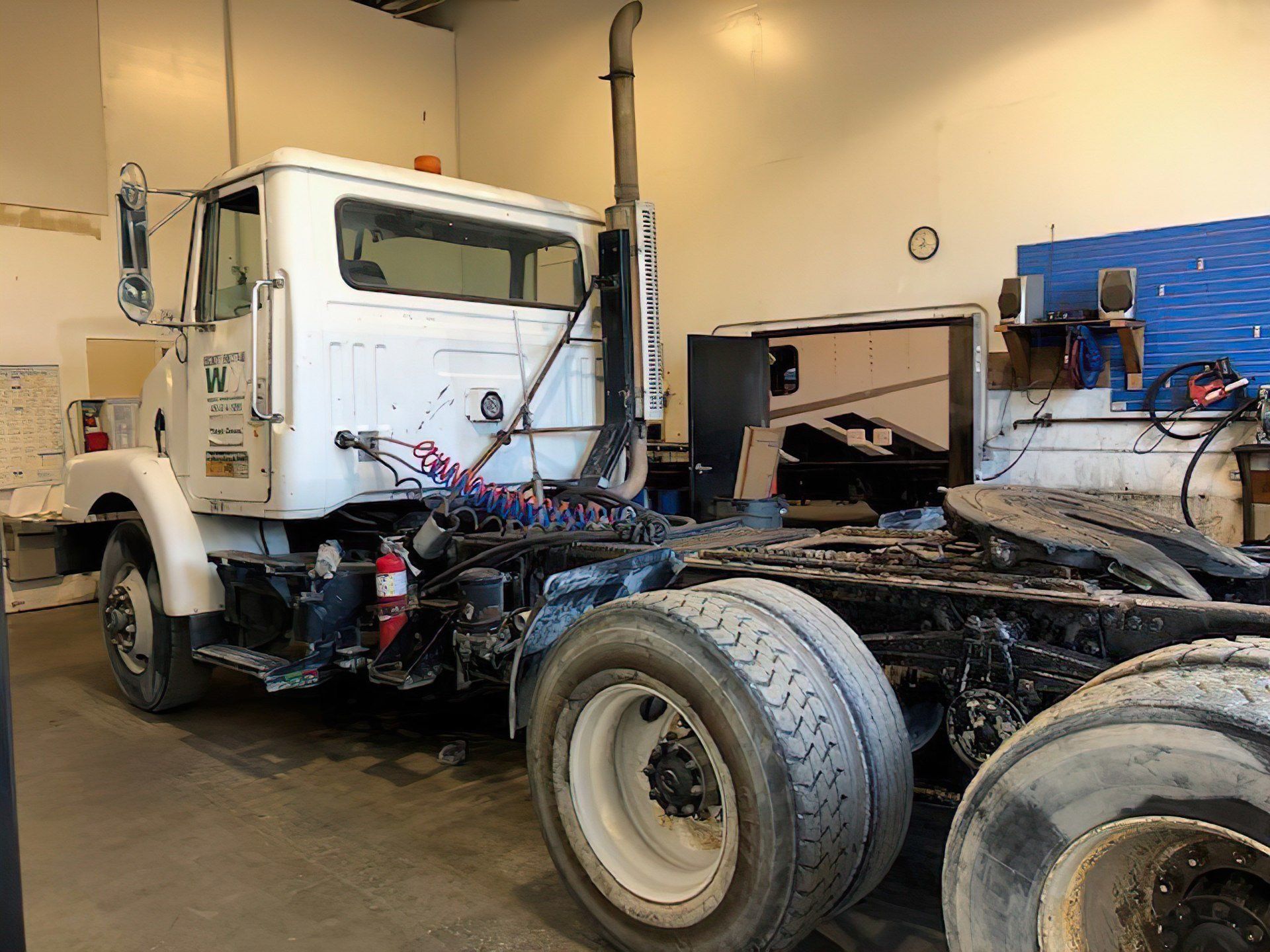 White semi-truck cab in a garage setting, with visible tires, air hoses, and smoke stack.