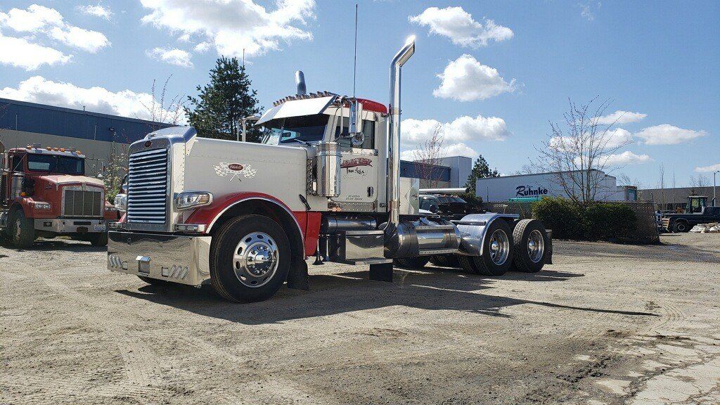 White and red semi-truck parked on dirt in a lot under a bright, partly cloudy sky.