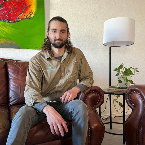 Man sitting on a brown leather couch, wearing tan shirt and gray pants. Beside him is a lamp and small table.