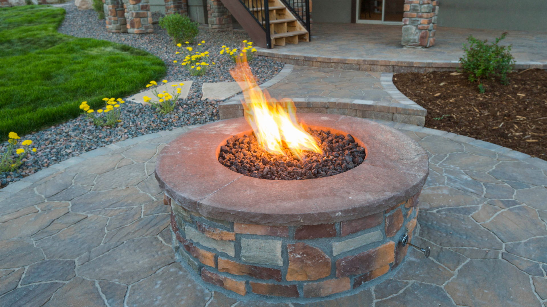 A round stone fire pit with flames, on a patio surrounded by landscaping.