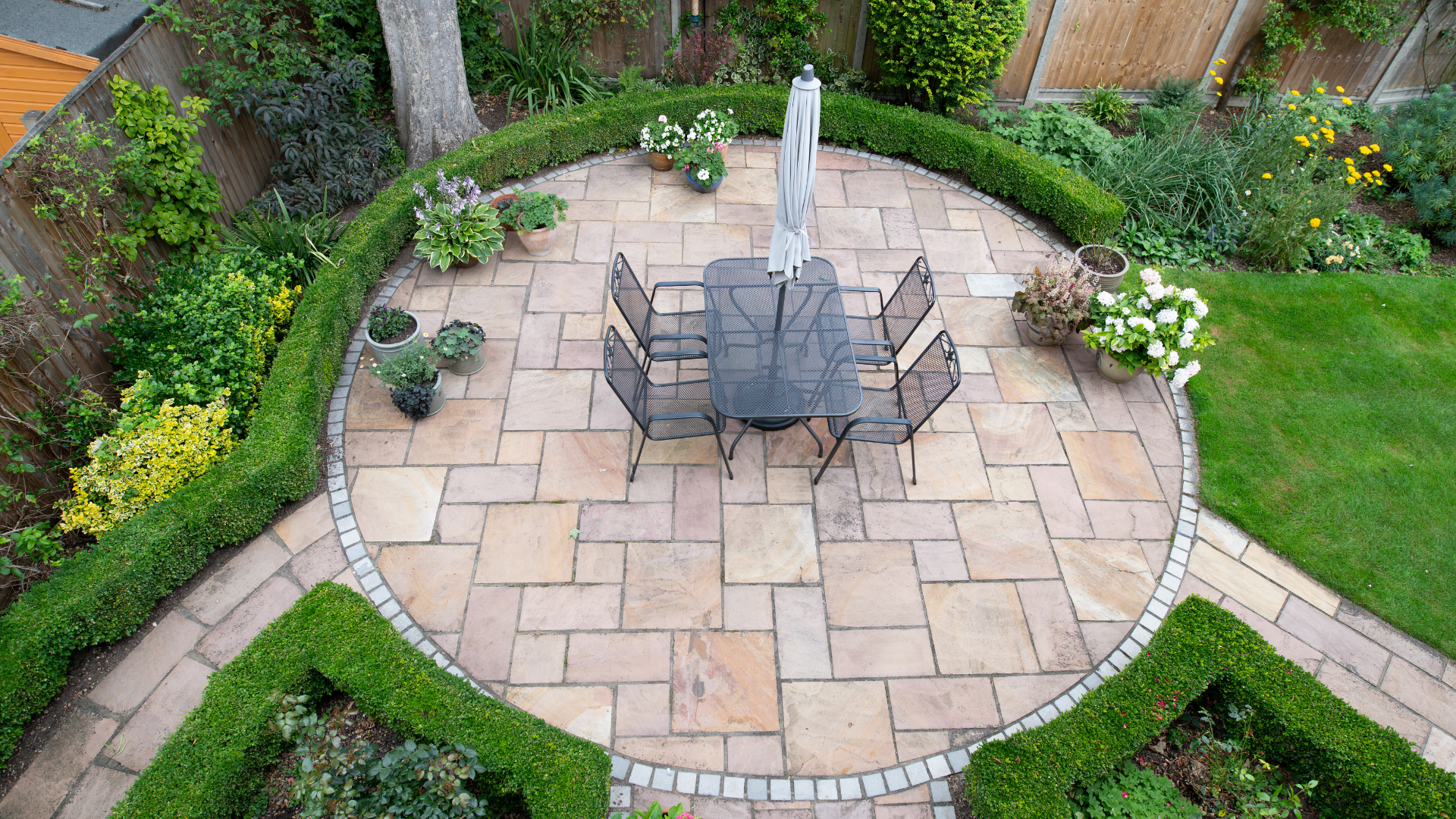 Overhead view of a circular patio with a table and chairs, surrounded by hedges and greenery.