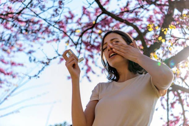 A woman holding an inhaler covering her nose with her hand while under a tree.