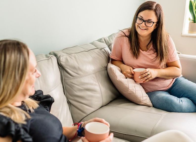 Two women are sitting on a couch talking and drinking coffee.