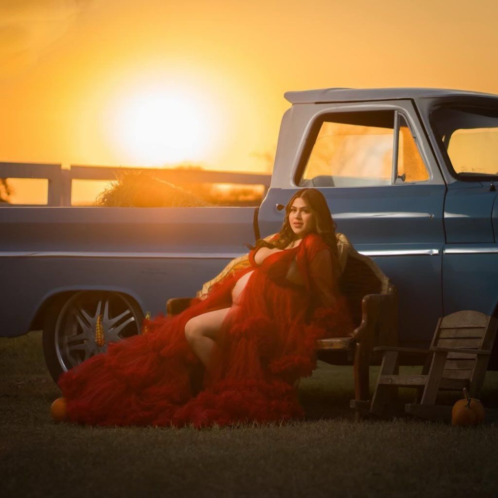 Pregnant person in red gown sits on truck, sunset background.