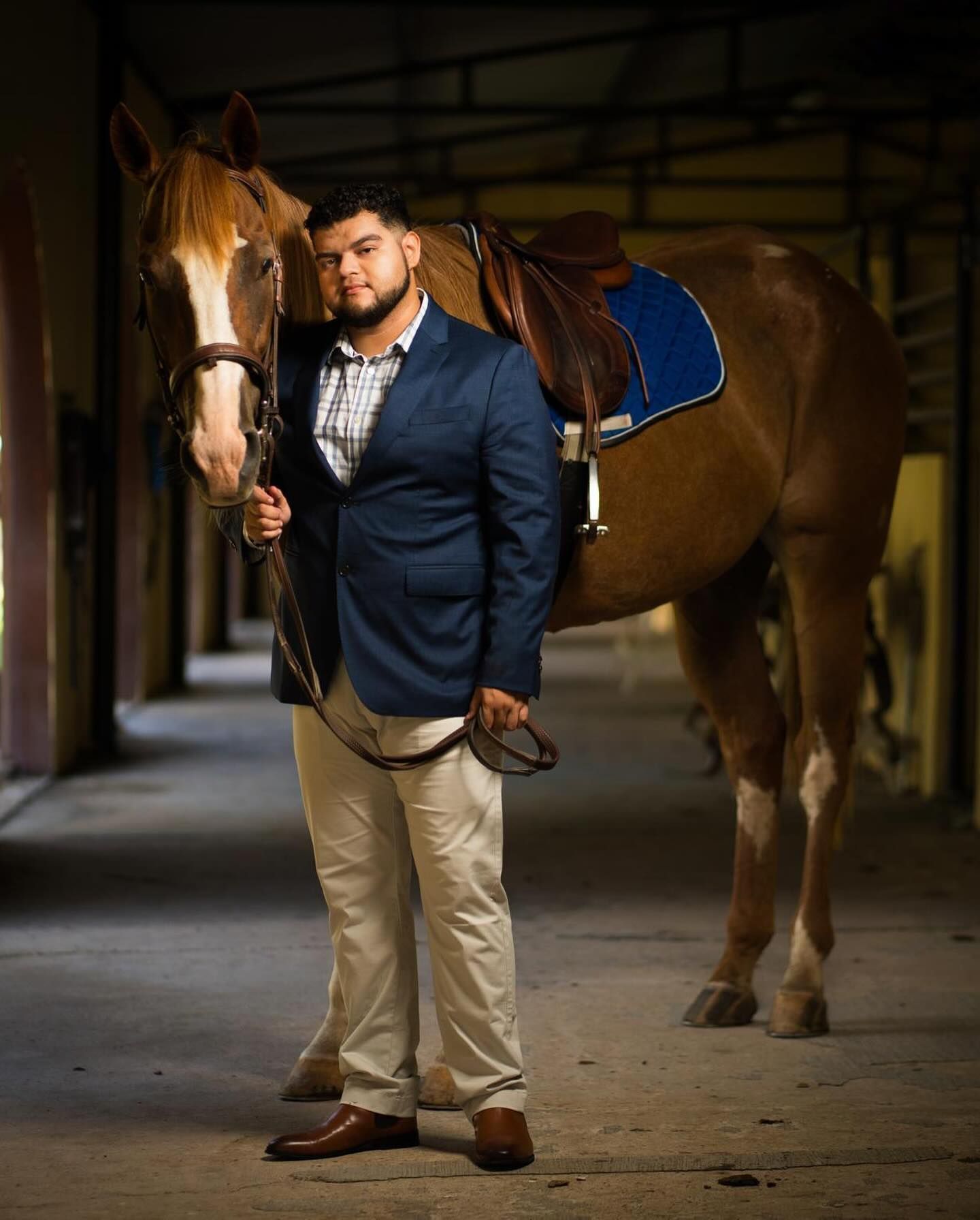 Man in blazer and khakis stands with a brown horse in a stable.