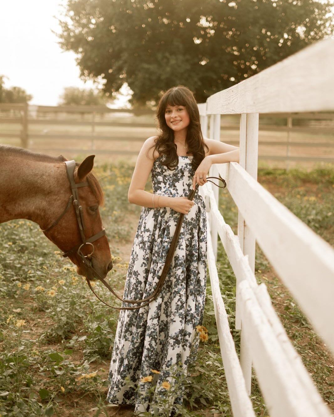 Woman in a floral dress leaning on a white fence, horse beside her in a field.