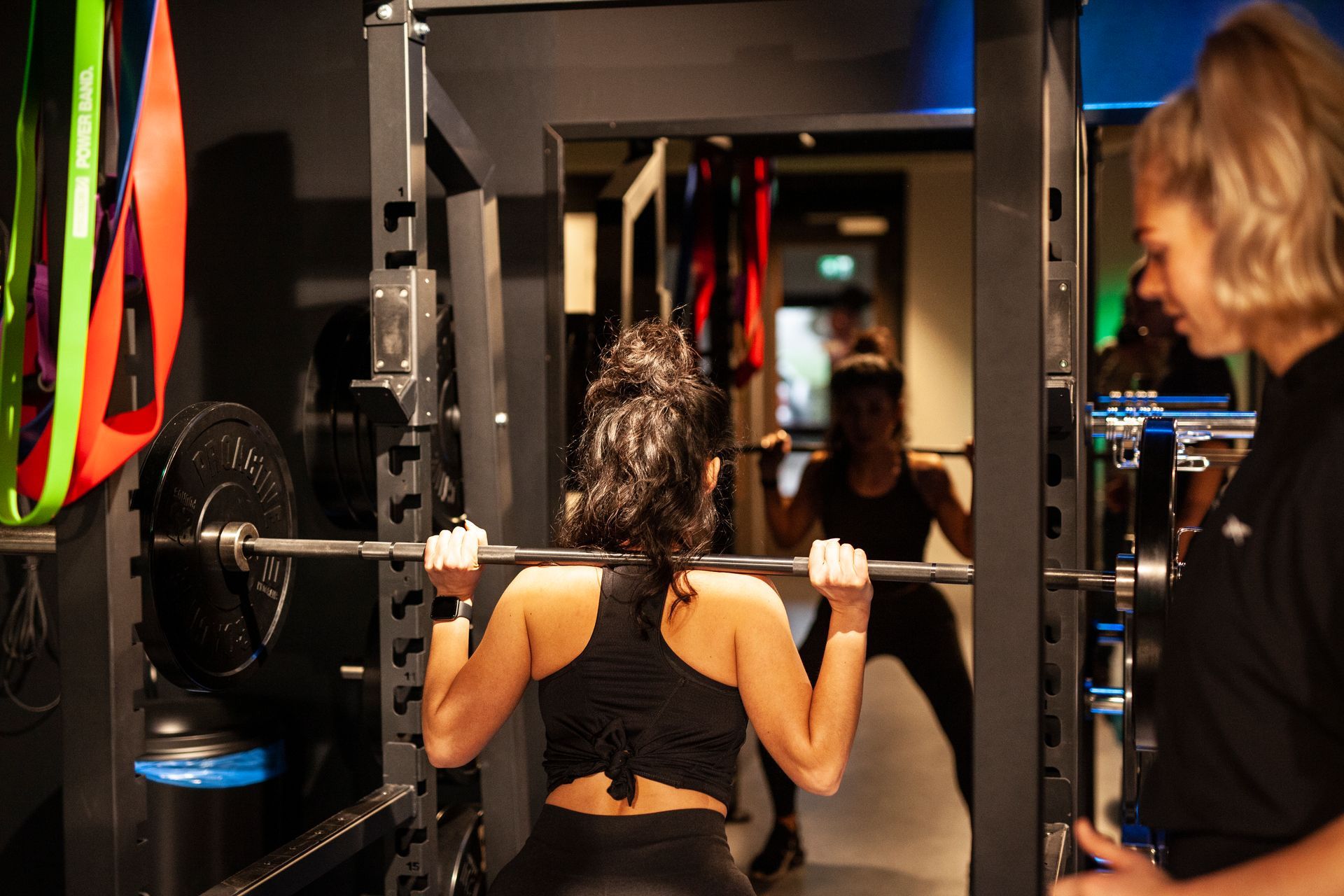 Woman performing a squat in a gym with a spotter. Black barbell, mirror, and equipment are visible.