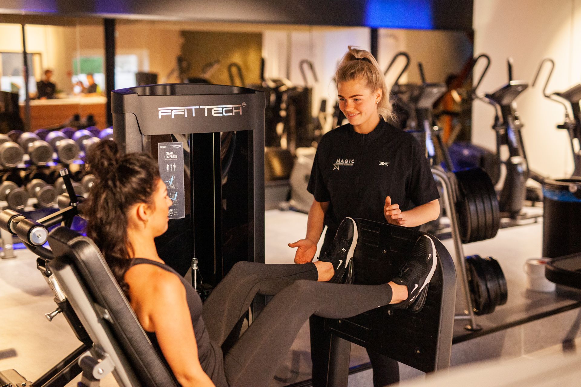 Een vrouw zit op een apparaat in de sportschool en praat met een andere vrouw.