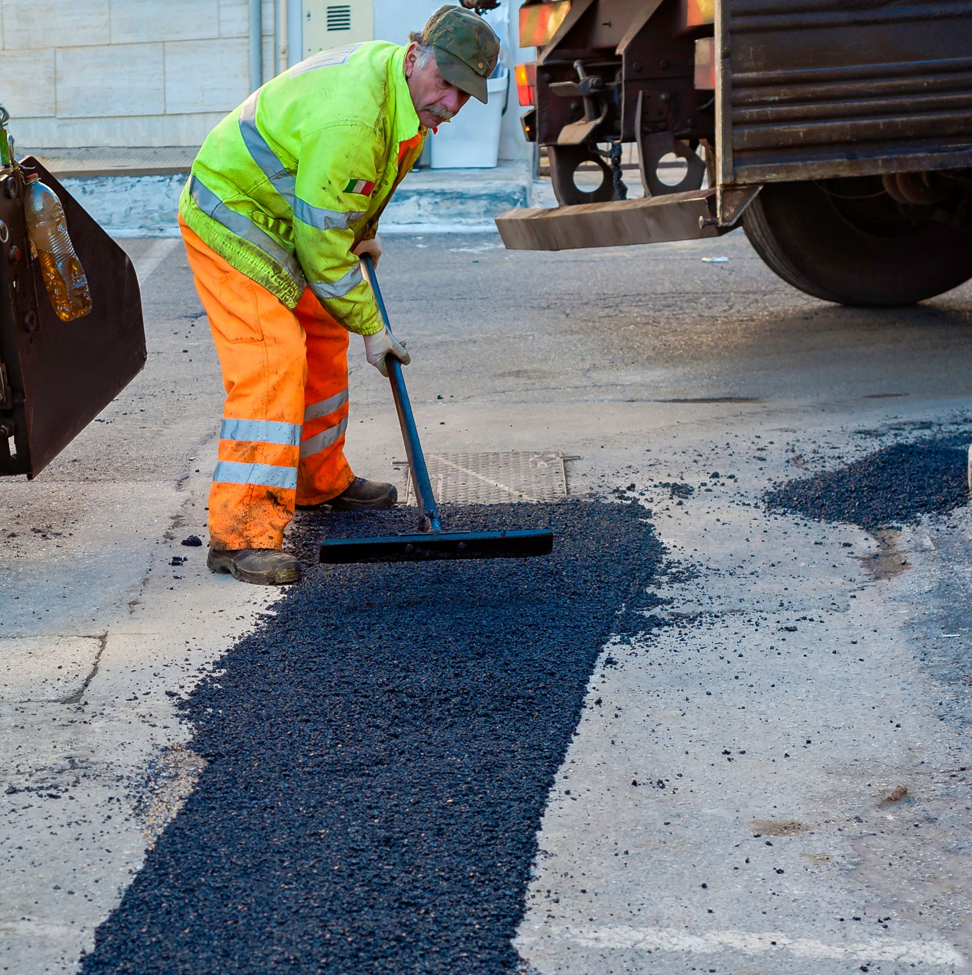 Road worker spreads asphalt with a squeegee on a road. Worker wears high-vis jacket and orange pants. Road worker spreads asphalt with a squeegee on a road. Worker wears high-vis jacket and orange pants.