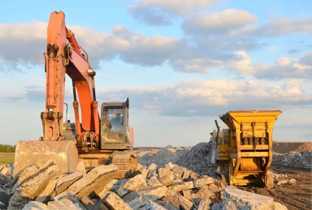 Orange excavator and yellow crusher on a pile of concrete, blue sky with clouds in the background.