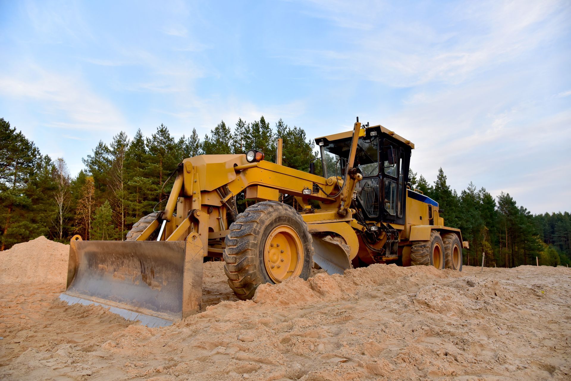 Yellow grader leveling sand on a construction site; trees and blue sky in background.