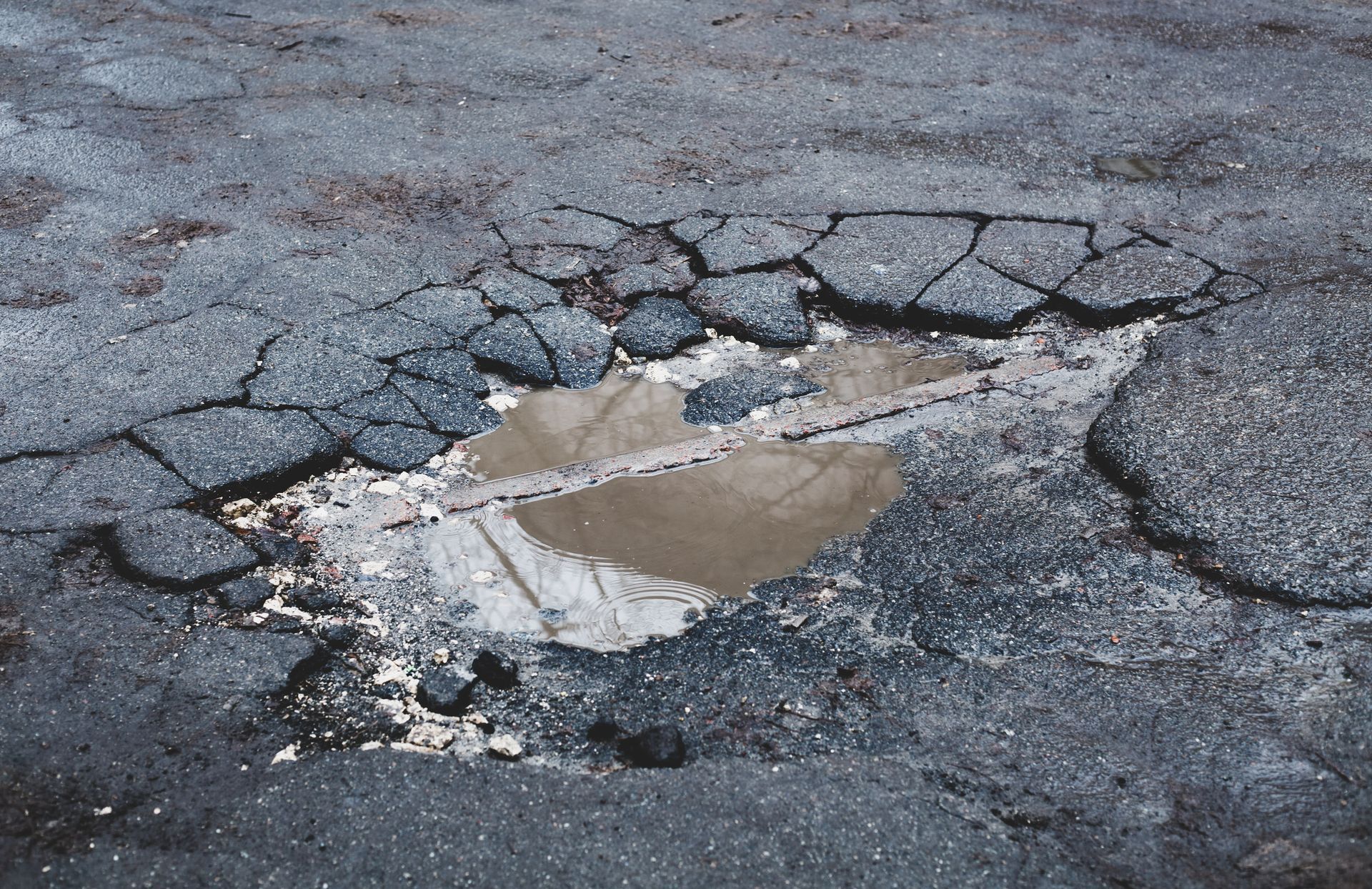Pothole in asphalt road, filled with water, surrounded by cracked pavement.