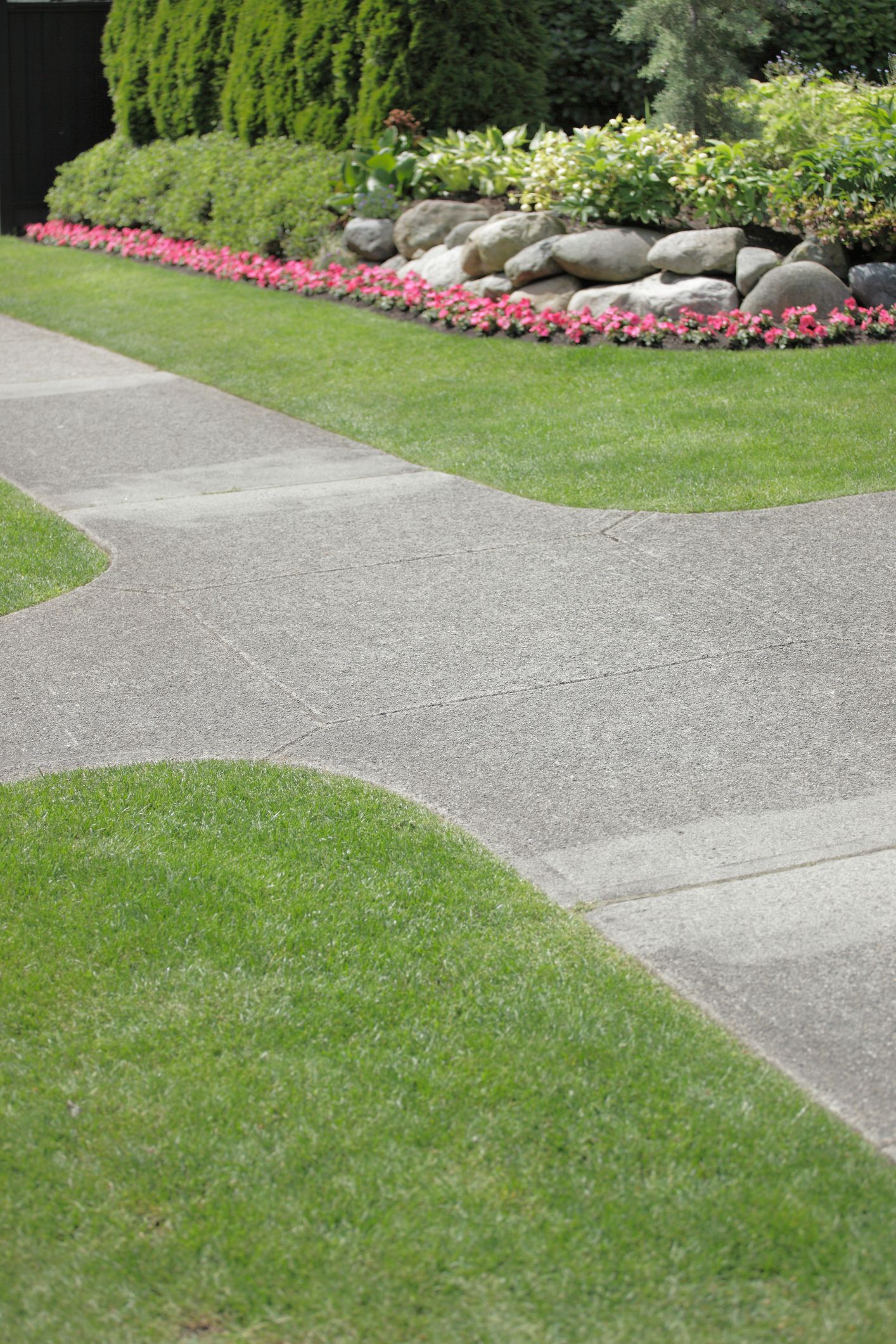 Gravel sidewalk curves through green lawn, leading toward a garden bed with pink flowers and large rocks.