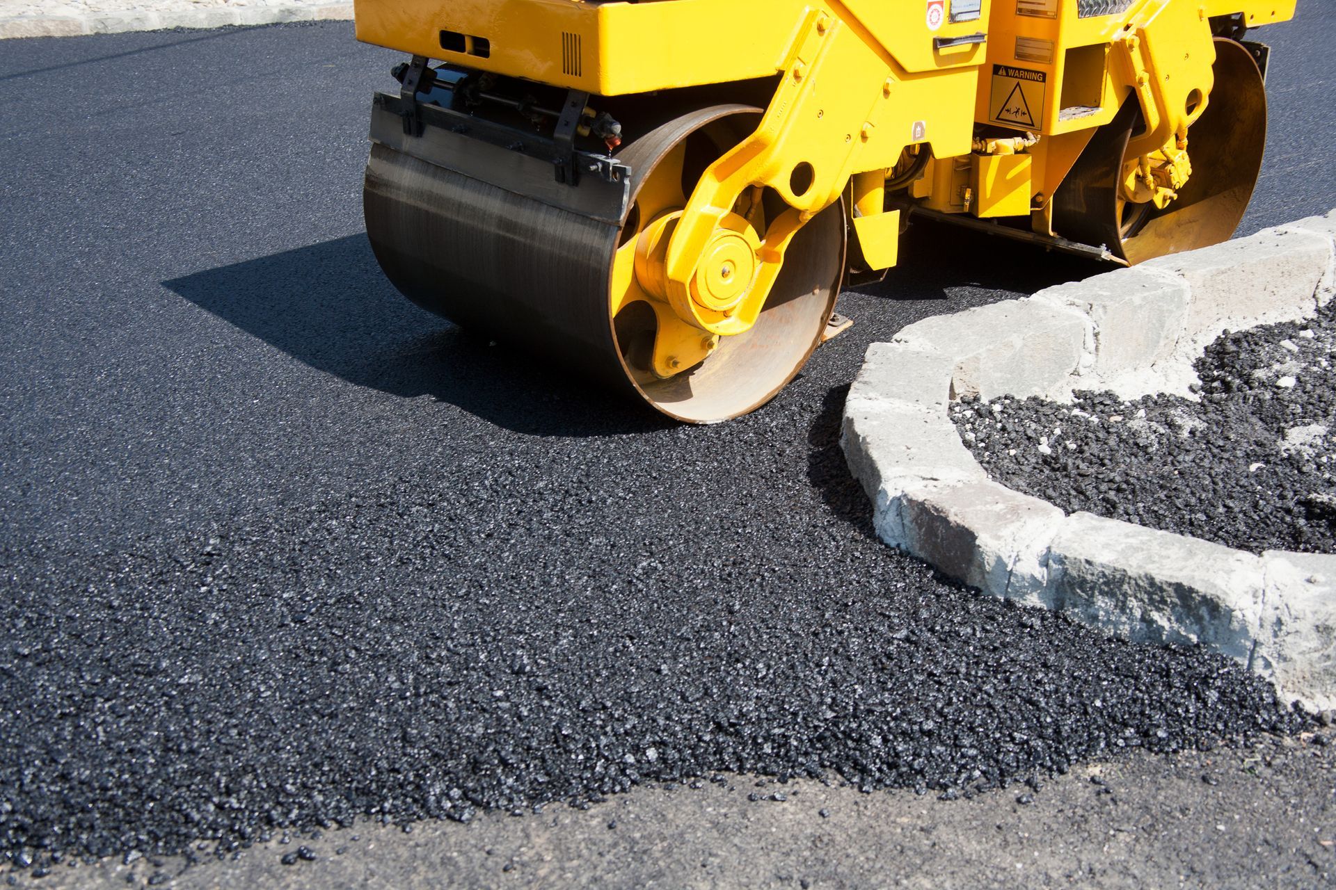 Yellow asphalt roller compacting fresh black asphalt next to a concrete curb.